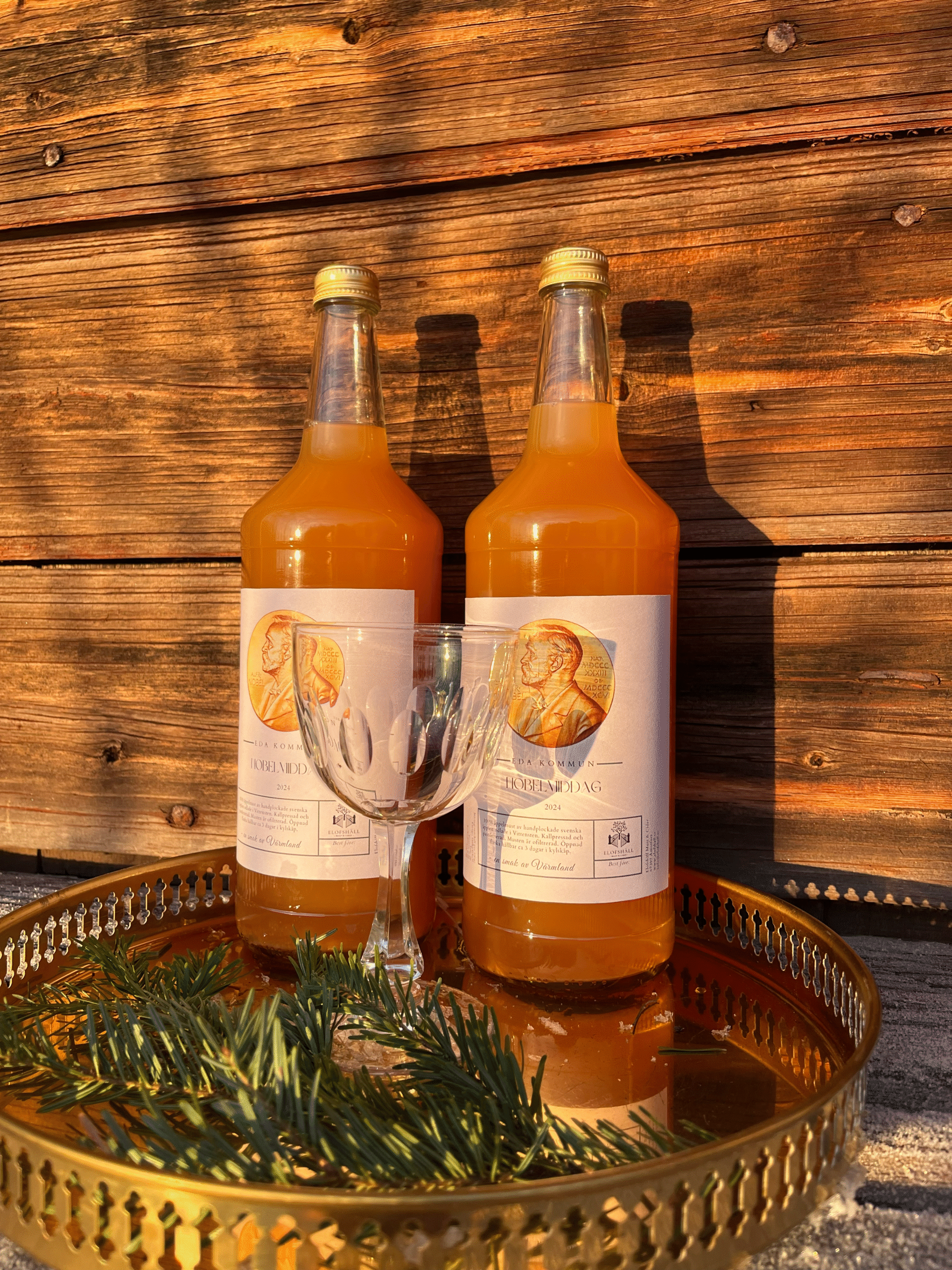 Två flaskor med orange dryck står på en gyllene bricka framför en trävägg i solsken. Framför flaskorna finns ett tomt vinglas och några gröna grankvistar. Ljuset skapar tydliga skuggor på väggen bakom. Scenen har en varm och rustik känsla.