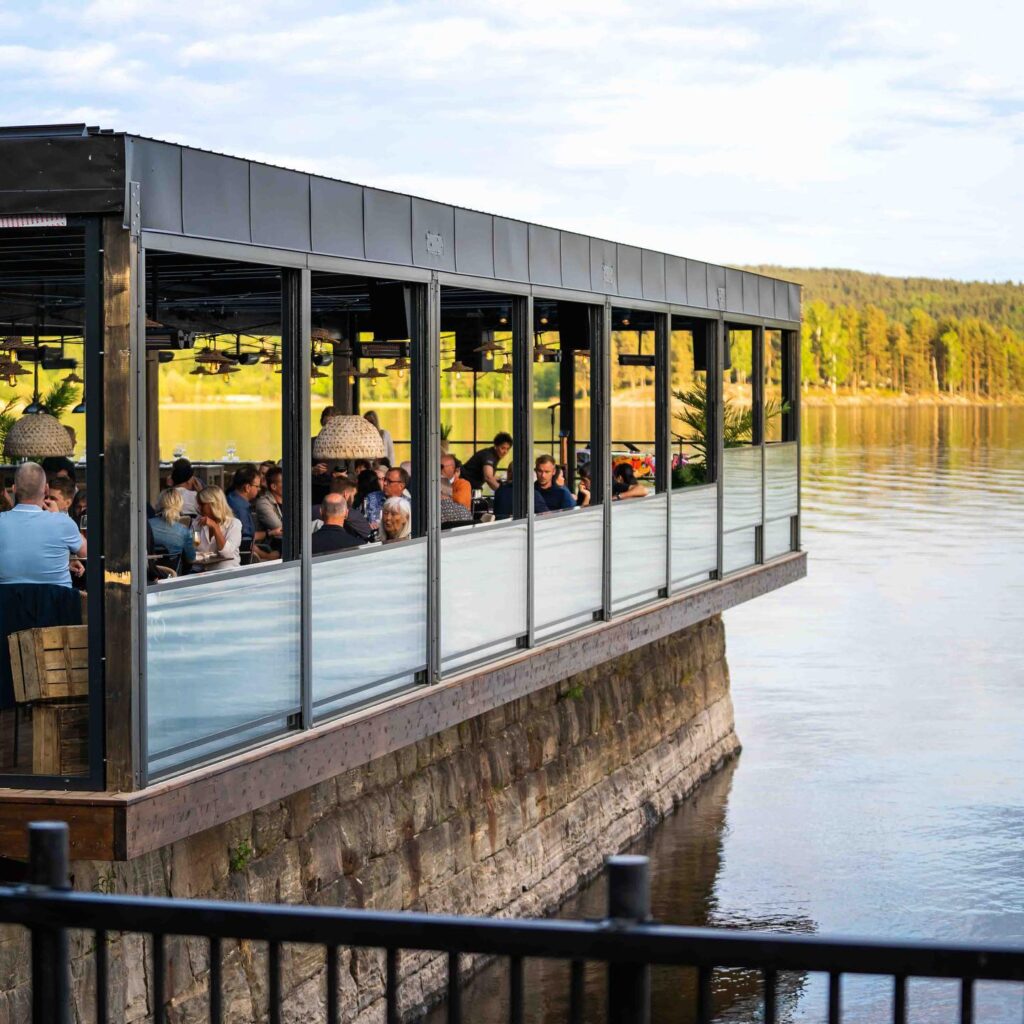 En inglasad restaurangterrass sträcker sig ut över vattnet med gäster sittande vid borden. Byggnaden vilar på en stenmur längs strandkanten, med lugnt vatten intill. I bakgrunden syns skogsklädda sluttningar i kvällsljus.