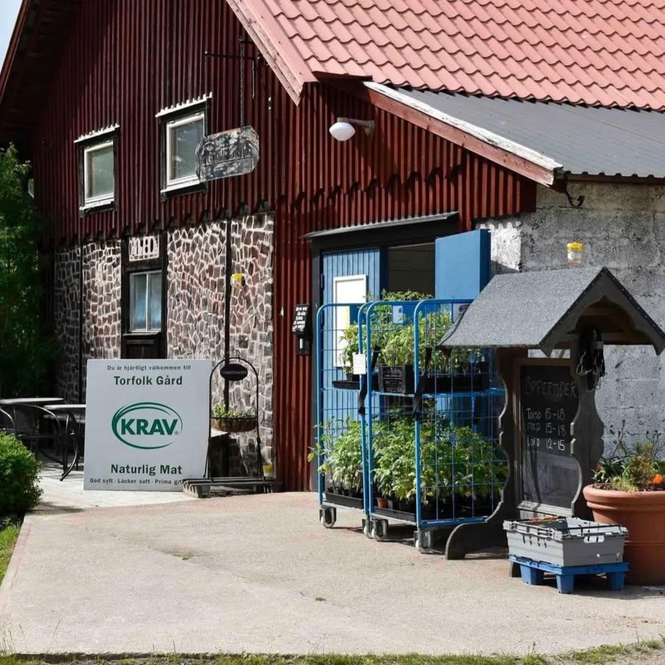 en röd träbyggnad med plåttak och stenfasad nedtill syns i en lantlig miljö. Utanför entrén står en skylt med texten “KRAV Naturlig Mat” samt vagnar med gröna plantor och odlingar. En mindre skylt vid dörren visar öppettider och ger intryck av gårdsbutik eller restaurang.