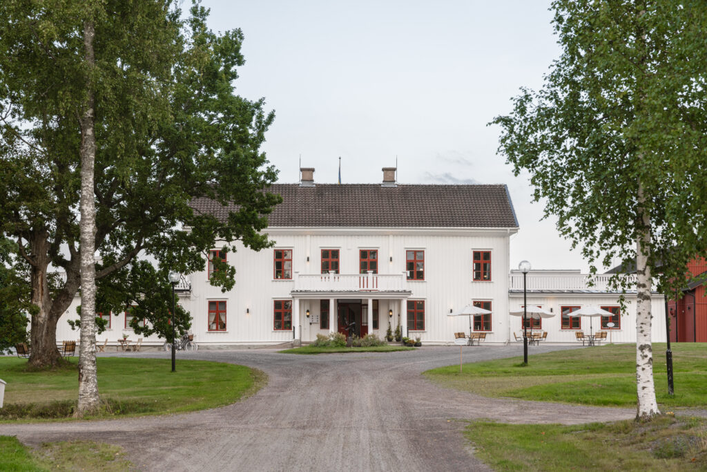 En vit herrgårdsbyggnad med röda fönster och mörkt tak ligger vid slutet av en grusväg. Byggnaden omges av gröna gräsmattor och höga träd på båda sidor. Uteservering med bord och parasoller syns framför huset.