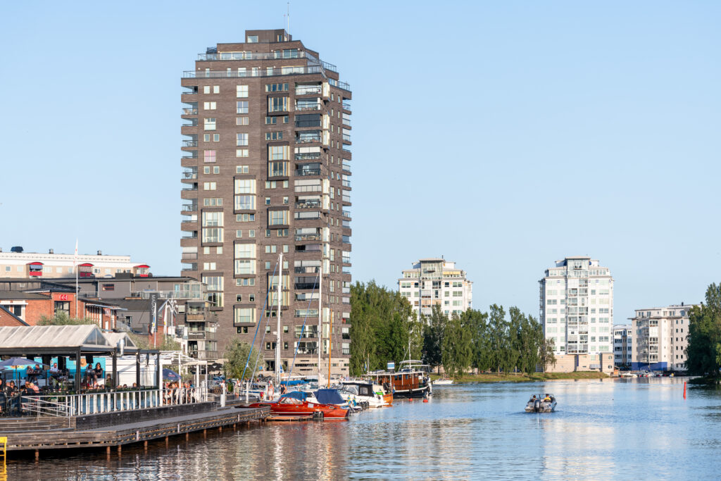 Ett högt flerbostadshus i mörkt tegel reser sig vid vattnet i en stadsmiljö. Längs kajen syns en uteservering, småbåtar och människor som rör sig vid bryggorna. I bakgrunden ligger fler ljusa bostadshus och grönska under en klar blå himmel.