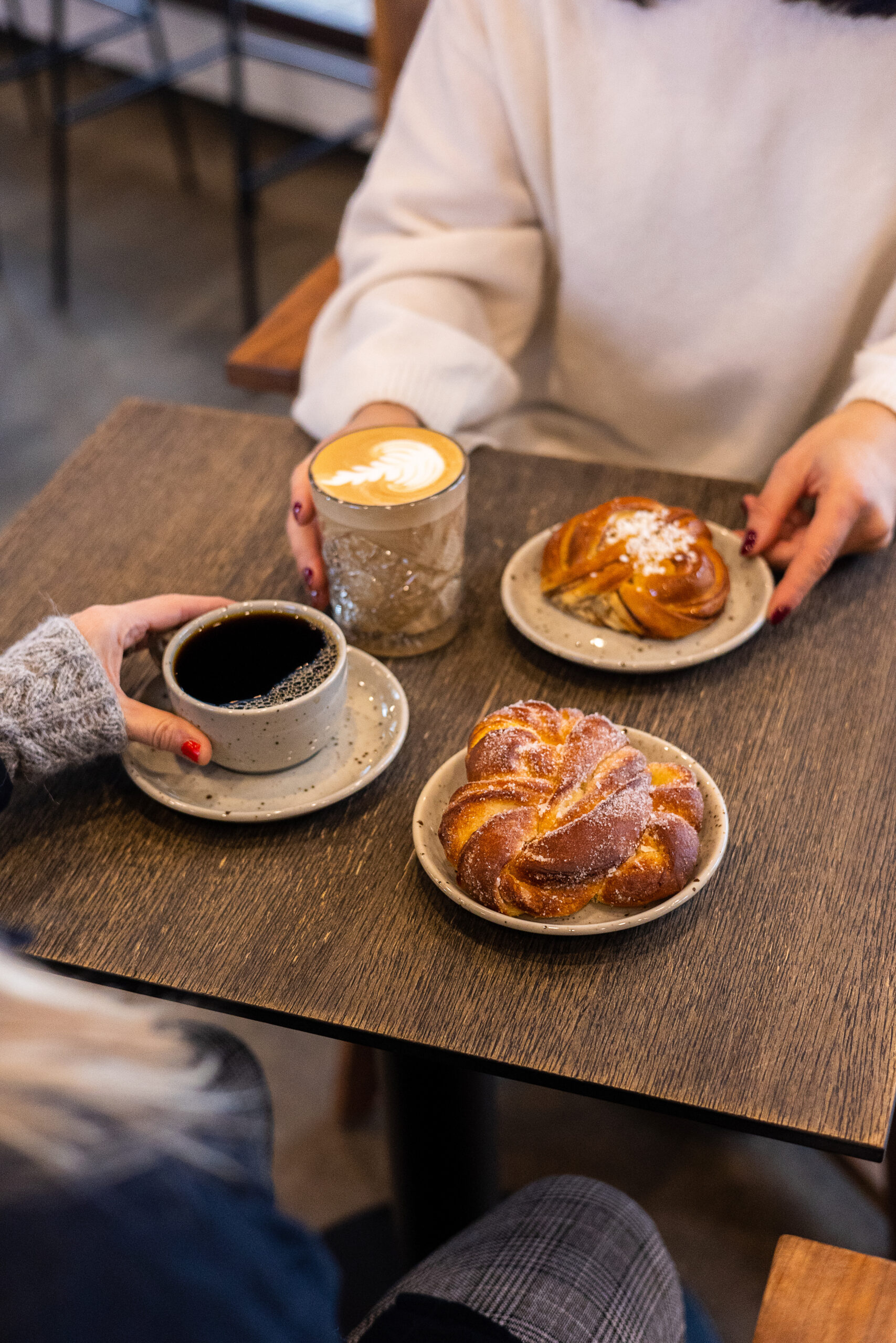 två personer sitter vid ett cafébord med kaffe och bakverk på små fat. 