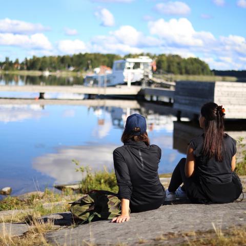 två kvinnor klädda i svarta kläder sitter på en klipphäll och tittar ut mot en båt som ligger förtöjd vid en brygga