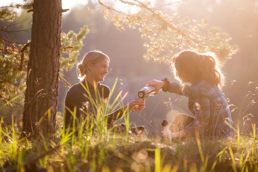 Two women sitting in a sunny forest drinking coffee.