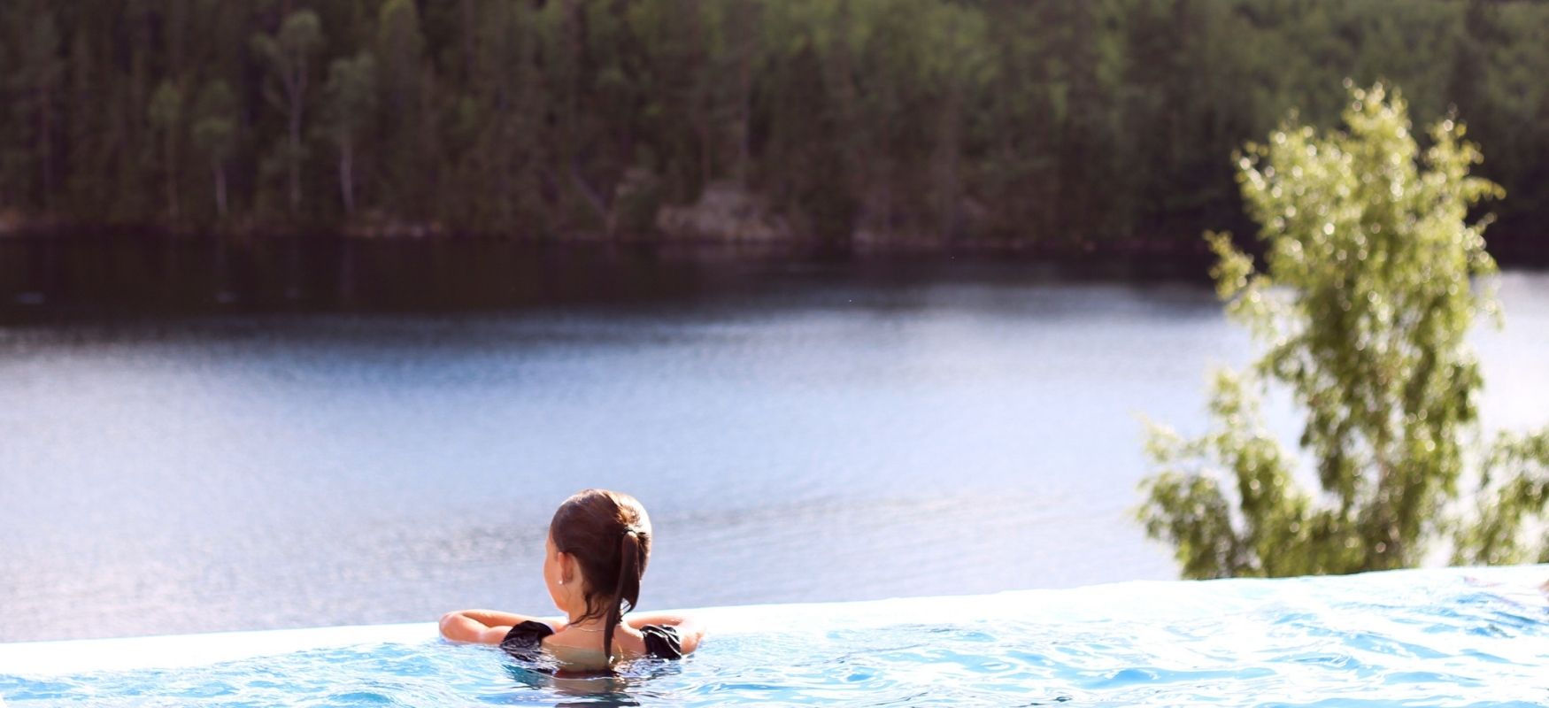 A girl swimming in a pool with a view over a lake and forest.